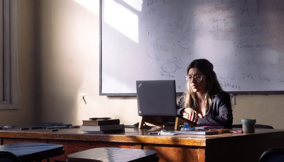 Profesora trabajando en su computador en un aula vacía, frente al tablero